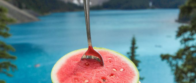 man holding watermelon with spoon inside in front of a lake.