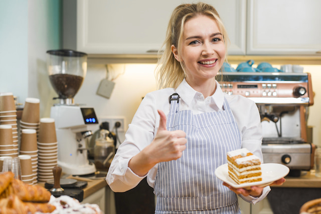 woman with thumbs up working in a café holding cake.